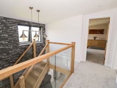 A wooden staircase with glass railing near a black tiled wall and a view into a bedroom with a wooden dresser and flowers at Phoenix House in Castlemorton