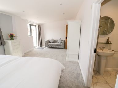 A bedroom with a white bed, white chest of drawers with flowers, gray sofa, white wardrobe, and an en suite bathroom with sink at Phoenix House in Castlemorton
