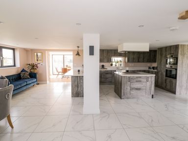 An open plan kitchen and living area with tiled floors a kitchen island grey cabinets a blue sofa and a dining area at Phoenix House in Castlemorton