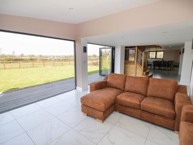 A living room with a brown sectional sofa tile flooring large open windows and a view of a fenced grassy yard at Phoenix House in Castlemorton