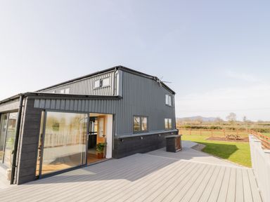 An exterior view of a modern building with sliding glass doors and a large deck overlooking a grassy field at Phoenix House in Castlemorton
