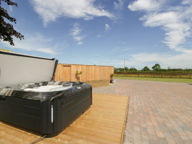 An outdoor hot tub on a wooden deck next to a paved area with a wooden fence and open sky at Phoenix House in Castlemorton