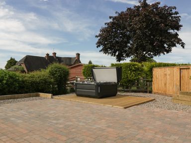 An outdoor area with a paved patio a wooden deck with a hot tub and surrounding hedges at Phoenix House in Castlemorton