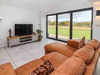 A living room with a brown leather sofa a TV on a wooden stand and large glass doors overlooking a garden at Phoenix House in Castlemorton