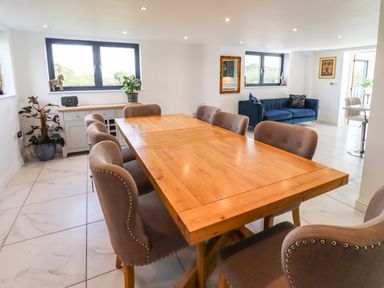 A dining area with a wooden table and upholstered chairs next to windows and a blue sofa at Phoenix House Castlemorton