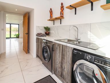A laundry room with a washing machine, dryer, sink, wooden shelves, and a potted plant at Phoenix House in Castlemorton