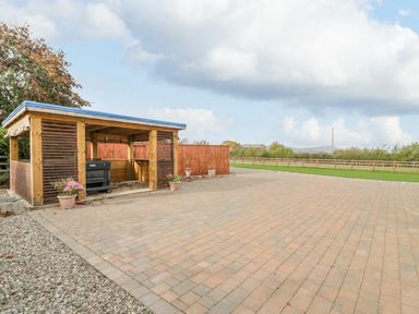 An outdoor wooden shelter with a stove and flower pots on a paved area near green grass and fenced land at Phoenix House in Castlemorton
