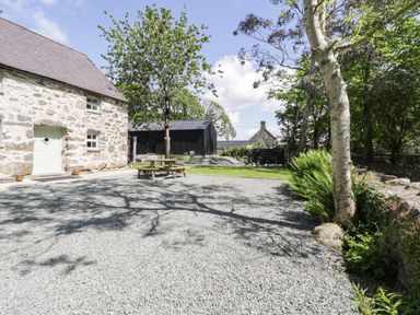 A stone building with a light door and windows next to a gravel area with a wooden picnic table surrounded by trees and plants at Stabla in Chwilog