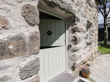 A half open pale green stable door set in a rustic stone wall with a diamond shaped window and a flower pot on the ground at Stabla in Chwilog