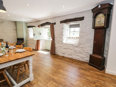A dining area with a wooden table set with plates and glasses next to an open door and windows with stone walls and a tall grandfather clock at Stabla in Chwilog