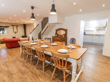 A dining area with a wooden table set with plates and glasses and wooden chairs next to a living area with a red sofa and a kitchen with white cabinets in the background at Stabla in Chwilog