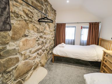 A bedroom with two single wooden beds a stone wall and windows with curtains at Stabla in Chwilog