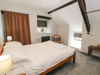 A bedroom with a double bed towels on the bed a chair with a cushion windows with blinds and exposed wooden beams at Stabla in Chwilog