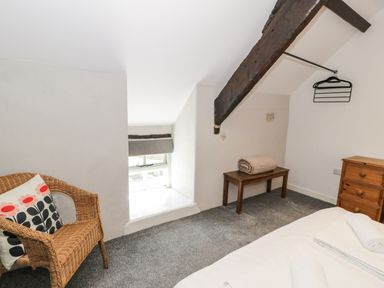 A bedroom with a wicker chair and cushion a wooden bench with a blanket a chest of drawers and a wooden beam on the ceiling at Stabla in Chwilog