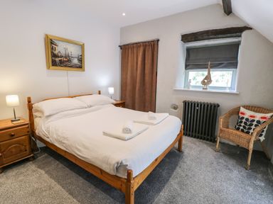A bedroom with a wooden bed with white linens two bedside tables with lamps a window with a gray blind and a wicker chair at Stabla in Chwilog