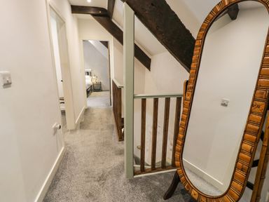 A hallway with carpeted floor leading to a bedroom with visible bed and lamp a wooden framed standing mirror and exposed ceiling beams at Stabla in Chwilog