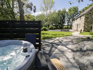 An outdoor hot tub next to a black wooden fence with a stone path leading to a stone building and picnic table at Stabla in Chwilog