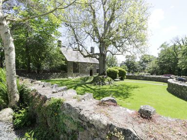 A stone building with a slate roof behind a stone wall and trees on a grassy lawn at Stabla in Chwilog