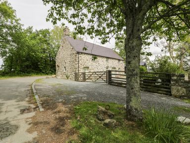 A stone house with a wooden gate and gravel driveway near a tree and a road at Stabla in Chwilog