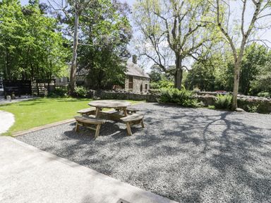An outdoor area with a round wooden picnic table and benches on gravel near trees and a stone building at Stabla in Chwilog