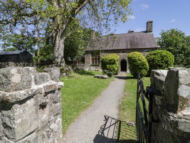 A stone house with a slate roof and arched doorway with a gravel path leading to an iron gate and stone pillars surrounded by grass and bushes at Stabla in Chwilog