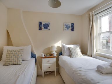 A bedroom with two single beds a nightstand with a lamp mugs and magazines and a window with curtains at The Ferryman in Weymouth