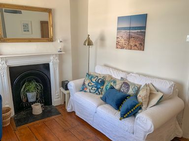 A white couch with decorative pillows in front of a wall with beach artwork beside a white fireplace with a plant inside at The Ferryman in Weymouth