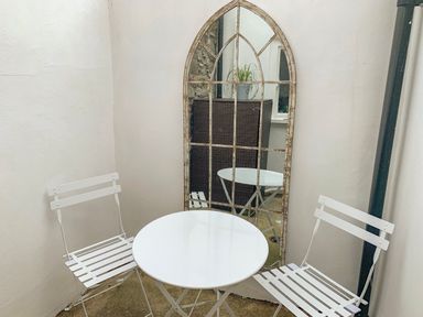 An outdoor seating area with two white metal chairs and a round white table in front of a tall arched mirror