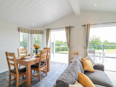 A dining area with a wooden table set for four and a gray sofa with pillows near large windows at Alder Lodge in Teigngrace near Newton Abbot