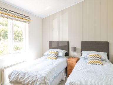 A bedroom with two single beds a bedside table with a lamp and a window with patterned blinds at Alder Lodge in Teigngrace near Newton Abbot