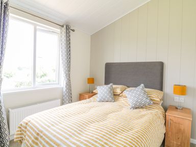 A bedroom with a bed with striped bedding two patterned cushions wooden bedside tables with lamps and curtains at Alder Lodge in Teigngrace near Newton Abbot