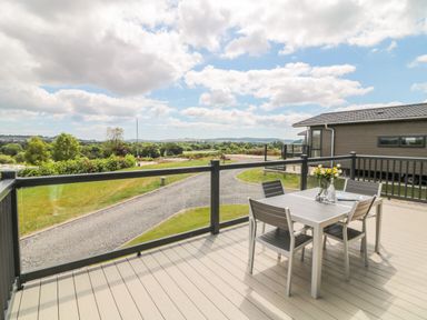 An outdoor deck with a table and four chairs overlooking a gravel driveway and grassy area at Birch Lodge in Teigngrace near Newton Abbot