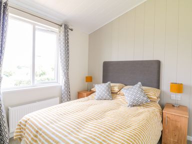 A bedroom with a bed with yellow and white striped bedding two wooden nightstands with lamps and patterned curtains at Birch Lodge in Teigngrace near Newton Abbot