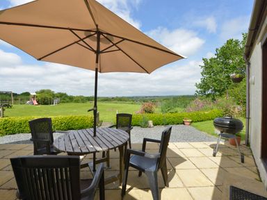 A patio with a round wooden table under a large umbrella and plastic chairs overlooking a grassy yard with playground equipment and a barbecue at Valley View in St Martin near Mawgan