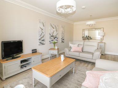 A living room with a sofa, coffee table, TV on a stand, side table with flowers, and wall art at Harbour Watch 2 in Weymouth