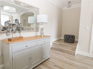A hallway with a grey sideboard with a lamp and decorative metal art and a mirror above it with wooden steps and a grey cushion at Harbour Watch 2 in Weymouth
