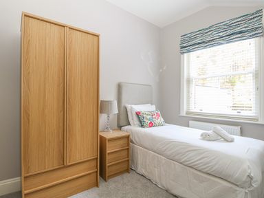 A bedroom with a single bed a wooden wardrobe a wooden bedside table with a lamp and a window with blinds and a patterned valance at Harbour Watch 2 Weymouth