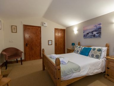 A bedroom with a wooden bed with pillows and rolled towels two wooden doors a brown chair and a bedside table with a lamp at Riverhouse Lodge in Upwey