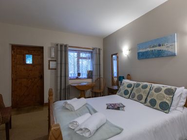 A bedroom with a bed towels and pillows a wooden door a window with curtains a table and chair a mirror and a painting at Riverhouse Lodge in Upwey
