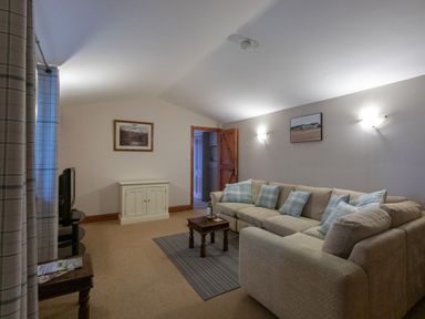 A living room with a beige sofa and blue and white pillows a small wooden coffee table on a rug a television on a stand a wooden side table with magazines curtains on a window and two framed pictures on walls at Riverhouse Lodge in Upwey