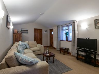 A living room with a beige sofa and blue cushions a wooden coffee table with wine glasses a television on a stand and a window with curtains at Riverhouse Lodge in Upwey