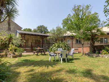 A garden with a wooden picnic table and chairs around a round table in front of a stone and beige building at Riverhouse Lodge in Upwey