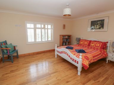A bedroom with a white bed with red and yellow bedding a wooden bookshelf a colorful chair and a window with white shutters at Yr Hen Ardd in Dale near Milford Haven