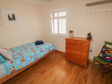 A small bedroom with a single bed with colorful bedding a wooden dresser a green chair with a decorative pillow and a window with white shutters at Yr Hen Ardd in Dale near Milford Haven