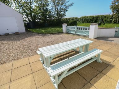 An outdoor area with a wooden picnic table and benches on a tiled patio next to a gravel driveway and grass with a gate and garage at Yr Hen Ardd in Dale near Milford Haven