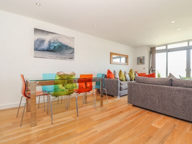 A dining area with a glass table and colorful chairs next to a living room with gray sofas and cushions at 8 Cribbar in Newquay