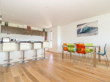 A kitchen with a breakfast bar with stools and a dining area with a glass table and colorful chairs at 8 Cribbar in Newquay