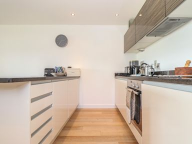 A modern kitchen with white cabinets gray countertops an oven with potholders a toaster a bread box and a clock on the wall at 8 Cribbar in Newquay