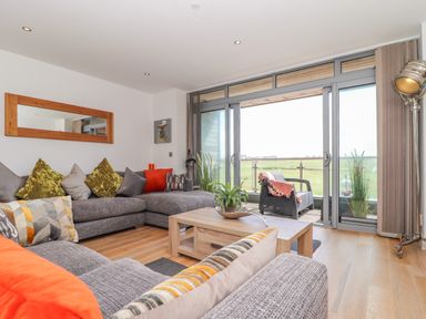 A living room with grey sofas and colorful cushions and a wooden coffee table with plant near sliding glass doors leading to a balcony with a chair at 8 Cribbar in Newquay