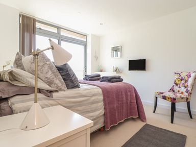 A bedroom with a bed with pillows and towels a side table with a lamp a wall mounted tv a decorative chair at 8 Cribbar in Newquay